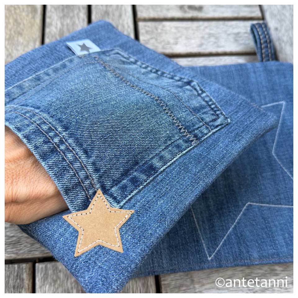 Eine Hand schlüpft in die Gesäßtasche eines blauen Upcycling-Topflappens aus Jeans, um die praktische Nutzung beim Greifen zu zeigen.