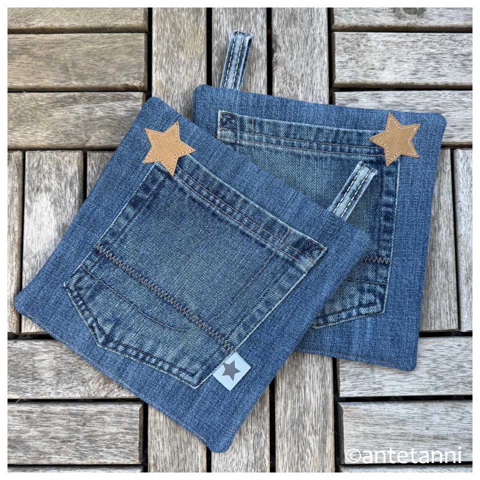 Zwei selbstgenähte, quadratische Topflappen aus blauem Jeansstoff mit aufgenähten Hosentaschen, kleinen SnapPap-Sternen und Gürtelschlaufen als Aufhänger auf einem Holzboden.
